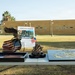 An outdoor display table set up for a Marine Spirit Monument event, featuring patriotic and military memorabilia. The table holds bronze sculptures, American flag items, and a USMC Eagle Globe and Anchor emblem plaque. A sign in the background reads 'Marine Spirit Monument' with a QR code, set against a military-style building.