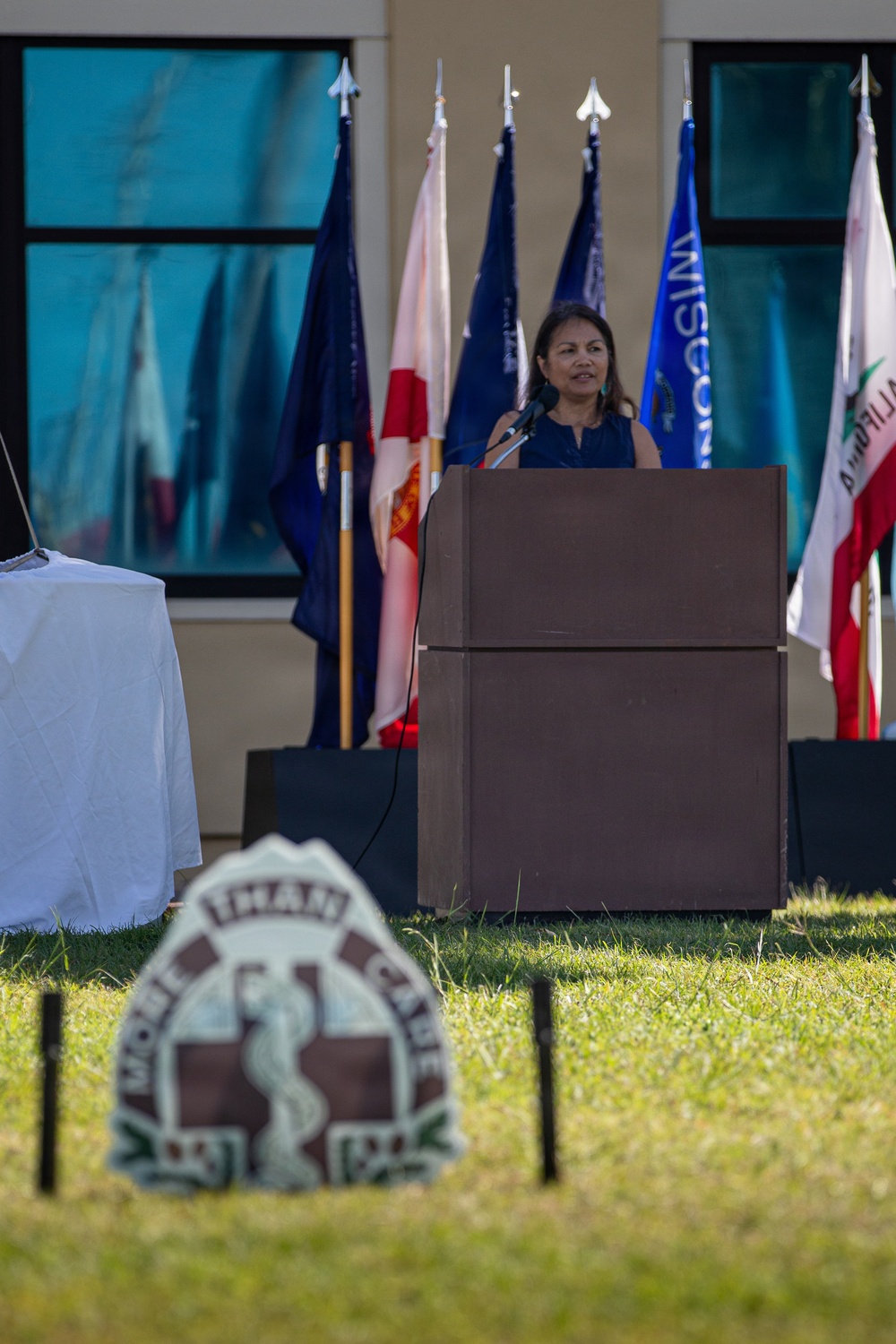 A woman speaks at a wooden podium during an outdoor ceremony or official event. Multiple state and organizational flags are displayed behind her, and a partially visible emblem or seal is in the foreground on the grass. The setting appears to be in front of a building with large windows.