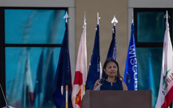 A woman speaks at a wooden podium during an outdoor ceremony or official event. Multiple state and organizational flags are displayed behind her, and a partially visible emblem or seal is in the foreground on the grass. The setting appears to be in front of a building with large windows.