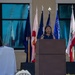 A woman speaks at a wooden podium during an outdoor ceremony or official event. Multiple state and organizational flags are displayed behind her, and a partially visible emblem or seal is in the foreground on the grass. The setting appears to be in front of a building with large windows.