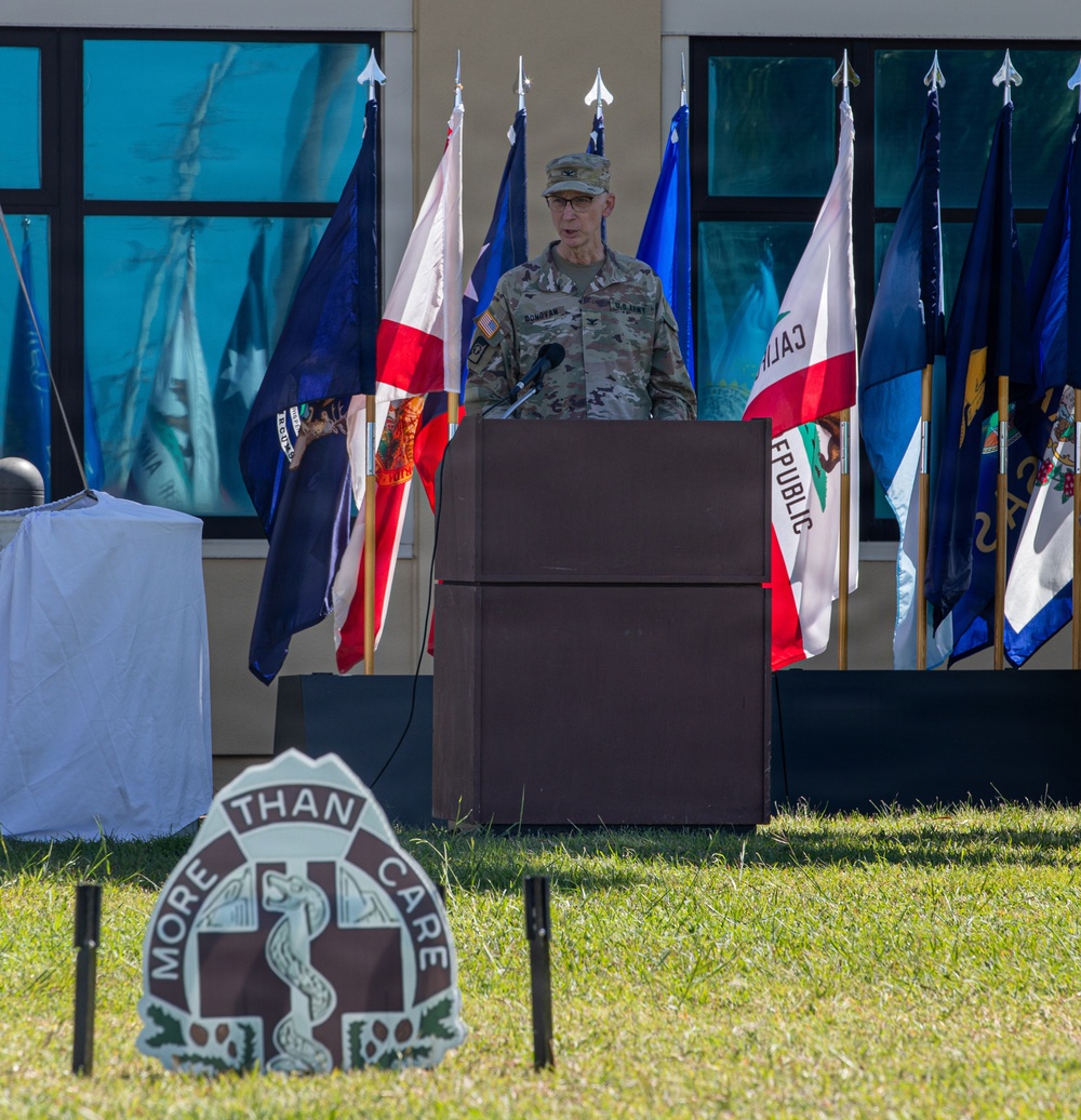 A U.S. Army officer in OCP (Operational Camouflage Pattern) uniform is speaking at a podium during an outdoor military ceremony. Multiple state and military flags are displayed behind the speaker. A sign in the foreground reads 'MORE THAN CARE' with a medical insignia, suggesting this is a U.S. Army Medical Command or military medical unit event.