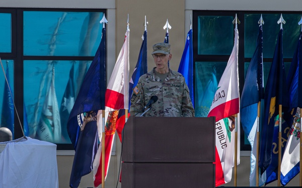 A U.S. Army officer in OCP (Operational Camouflage Pattern) uniform is speaking at a podium during an outdoor military ceremony. Multiple state and military flags are displayed behind the speaker. A sign in the foreground reads 'MORE THAN CARE' with a medical insignia, suggesting this is a U.S. Army Medical Command or military medical unit event.