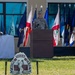 A U.S. Army officer in OCP (Operational Camouflage Pattern) uniform is speaking at a podium during an outdoor military ceremony. Multiple state and military flags are displayed behind the speaker. A sign in the foreground reads 'MORE THAN CARE' with a medical insignia, suggesting this is a U.S. Army Medical Command or military medical unit event.