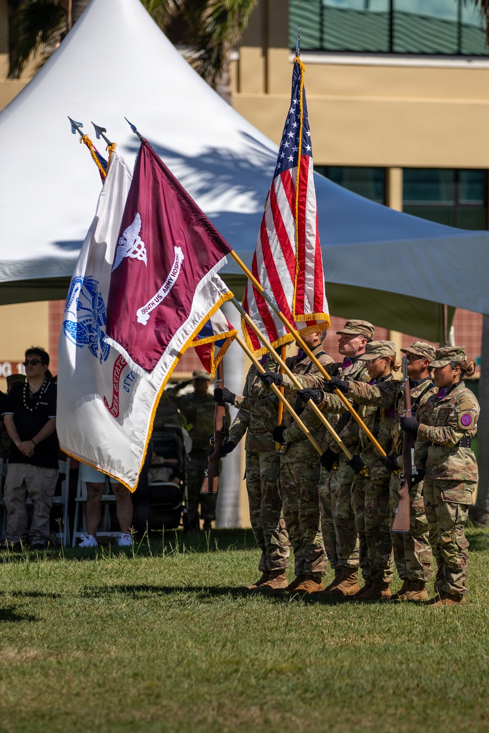 A U.S. Army color guard presents flags during a formal military ceremony outdoors. Soldiers in Army Combat Uniforms (ACU/OCP pattern) carry multiple flags including the U.S. national flag and what appears to be the 1994th U.S. Army Hospital guidon. The ceremony takes place on a grassy area with a white tent canopy and building visible in the background.