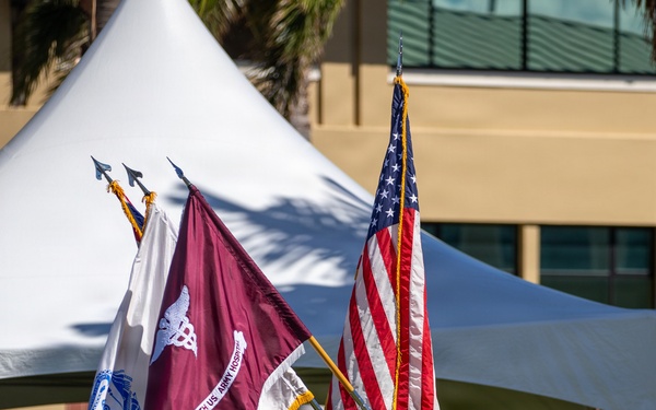 A U.S. Army color guard presents flags during a formal military ceremony outdoors. Soldiers in Army Combat Uniforms (ACU/OCP pattern) carry multiple flags including the U.S. national flag and what appears to be the 1994th U.S. Army Hospital guidon. The ceremony takes place on a grassy area with a white tent canopy and building visible in the background.