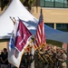 A U.S. Army color guard presents flags during a formal military ceremony outdoors. Soldiers in Army Combat Uniforms (ACU/OCP pattern) carry multiple flags including the U.S. national flag and what appears to be the 1994th U.S. Army Hospital guidon. The ceremony takes place on a grassy area with a white tent canopy and building visible in the background.