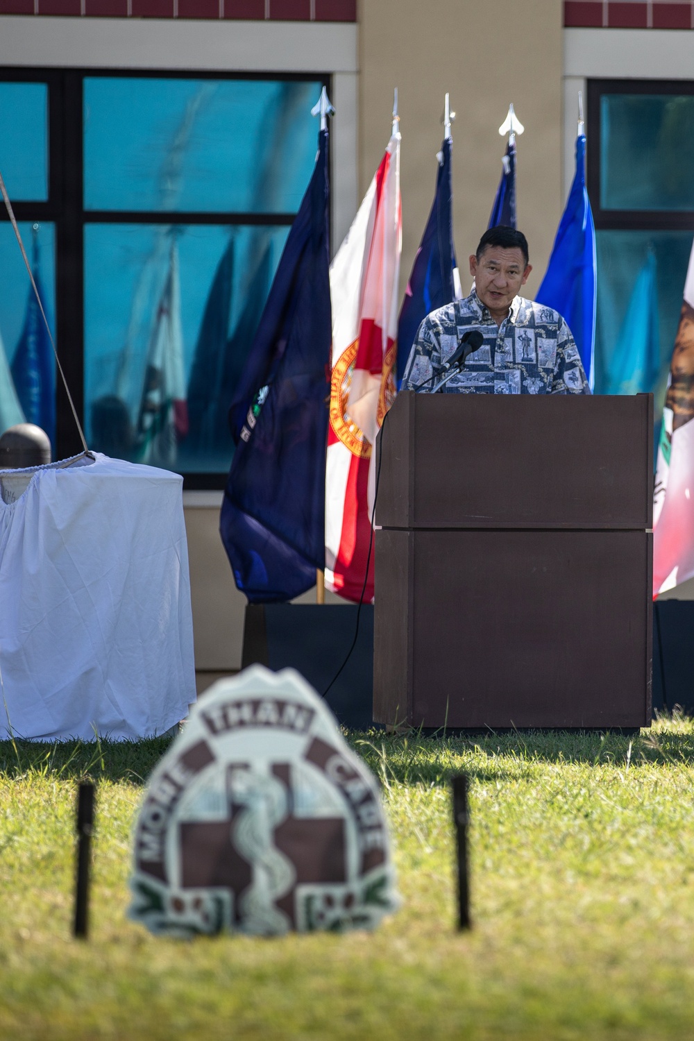 A man in a patterned Hawaiian-style shirt speaks at a podium during what appears to be an outdoor military or official ceremony. Multiple flags are displayed behind him, and a partially covered object is visible to the left, suggesting an unveiling event. In the foreground, a sign or emblem with the text 'MORE THAN CAPABLE' is visible on the grass.