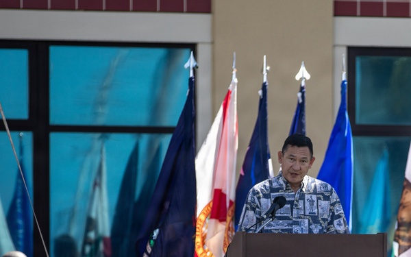 A man in a patterned Hawaiian-style shirt speaks at a podium during what appears to be an outdoor military or official ceremony. Multiple flags are displayed behind him, and a partially covered object is visible to the left, suggesting an unveiling event. In the foreground, a sign or emblem with the text 'MORE THAN CAPABLE' is visible on the grass.
