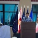 A man in a patterned Hawaiian-style shirt speaks at a podium during what appears to be an outdoor military or official ceremony. Multiple flags are displayed behind him, and a partially covered object is visible to the left, suggesting an unveiling event. In the foreground, a sign or emblem with the text 'MORE THAN CAPABLE' is visible on the grass.