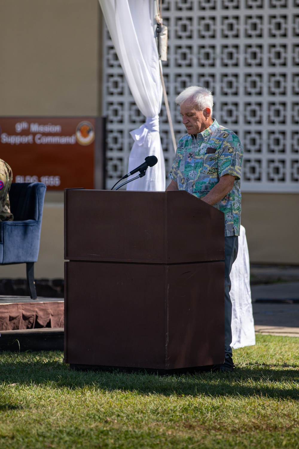 An elderly man with white hair stands at a wooden podium with a microphone, appearing to speak or read at an outdoor military ceremony. A sign in the background reads '9th Mission Support Command, BLDG 1555,' indicating this is a U.S. Army installation event. The setting includes decorative white draping, a latticed wall, and a manicured grass lawn.
