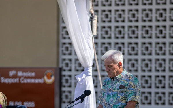 An elderly man with white hair stands at a wooden podium with a microphone, appearing to speak or read at an outdoor military ceremony. A sign in the background reads '9th Mission Support Command, BLDG 1555,' indicating this is a U.S. Army installation event. The setting includes decorative white draping, a latticed wall, and a manicured grass lawn.
