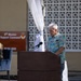 An elderly man with white hair stands at a wooden podium with a microphone, appearing to speak or read at an outdoor military ceremony. A sign in the background reads '9th Mission Support Command, BLDG 1555,' indicating this is a U.S. Army installation event. The setting includes decorative white draping, a latticed wall, and a manicured grass lawn.