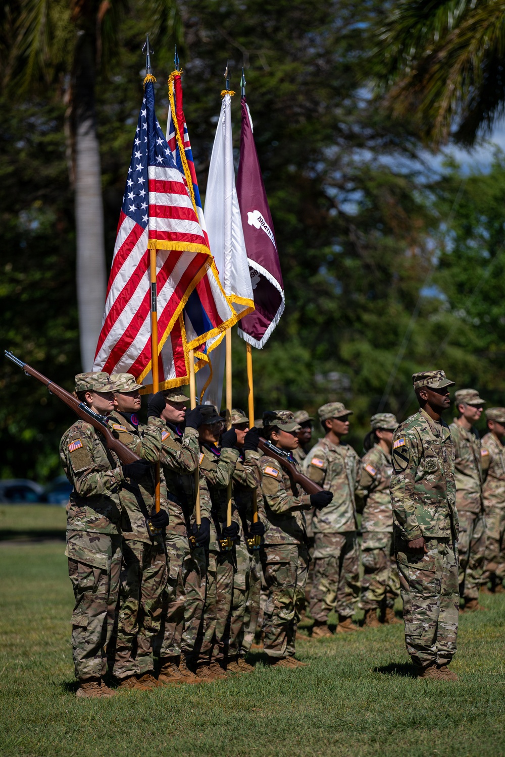A U.S. Army color guard and formation stand at attention during a military ceremony, likely on a base in Hawaii given the tropical vegetation and presence of the Hawaii state flag. Soldiers in OCP (Operational Camouflage Pattern) uniforms carry multiple flags including the U.S. national flag, the Hawaii state flag, a white Army flag, and a maroon unit guidon. The ceremony appears to be a formal military event such as a change of command, activation, or unit ceremony.