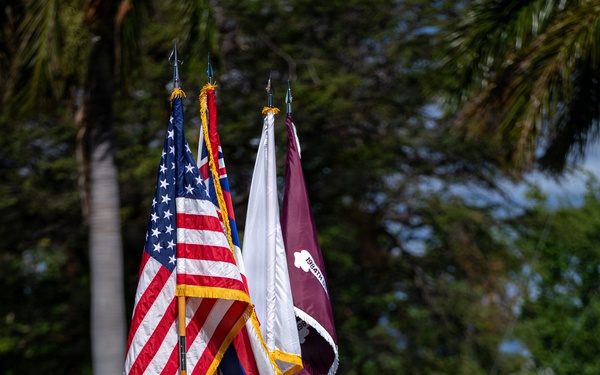 A U.S. Army color guard and formation stand at attention during a military ceremony, likely on a base in Hawaii given the tropical vegetation and presence of the Hawaii state flag. Soldiers in OCP (Operational Camouflage Pattern) uniforms carry multiple flags including the U.S. national flag, the Hawaii state flag, a white Army flag, and a maroon unit guidon. The ceremony appears to be a formal military event such as a change of command, activation, or unit ceremony.