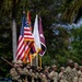 A U.S. Army color guard and formation stand at attention during a military ceremony, likely on a base in Hawaii given the tropical vegetation and presence of the Hawaii state flag. Soldiers in OCP (Operational Camouflage Pattern) uniforms carry multiple flags including the U.S. national flag, the Hawaii state flag, a white Army flag, and a maroon unit guidon. The ceremony appears to be a formal military event such as a change of command, activation, or unit ceremony.
