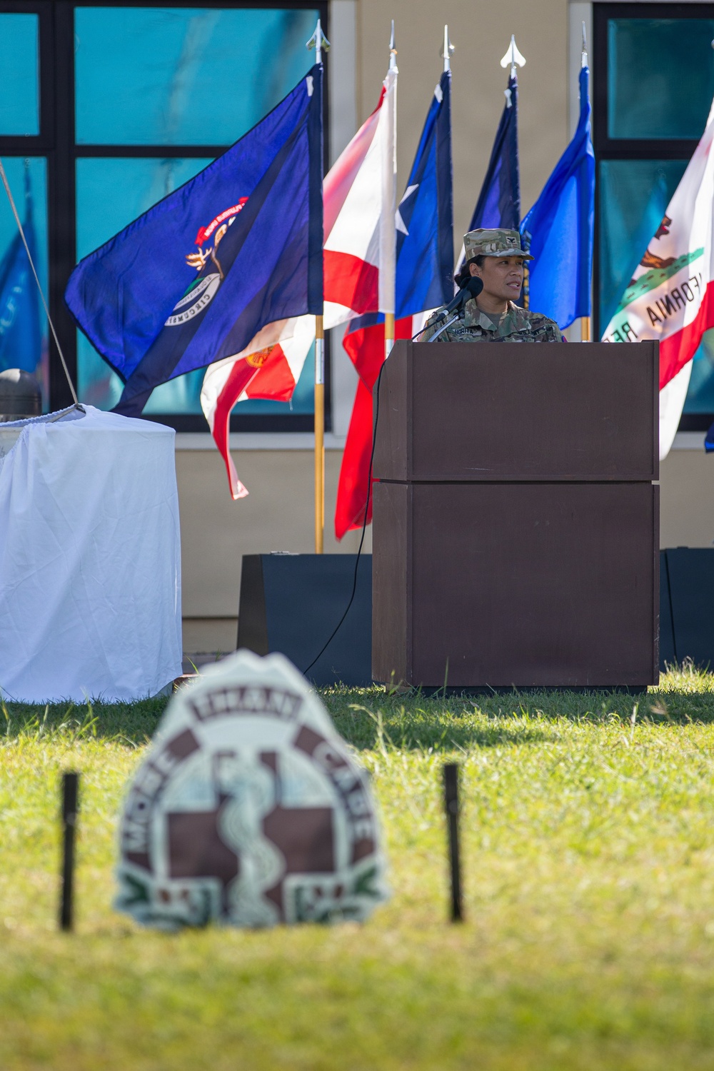 A military service member in camouflage uniform and patrol cap stands at a wooden podium with a microphone, appearing to deliver a speech at an outdoor military ceremony. Multiple U.S. state flags are displayed prominently in the background, including identifiable flags of Louisiana, Texas, and California. In the foreground, an out-of-focus military insignia sign is visible on the grass, consistent with a U.S. Army Medical Command or similar medical unit emblem.