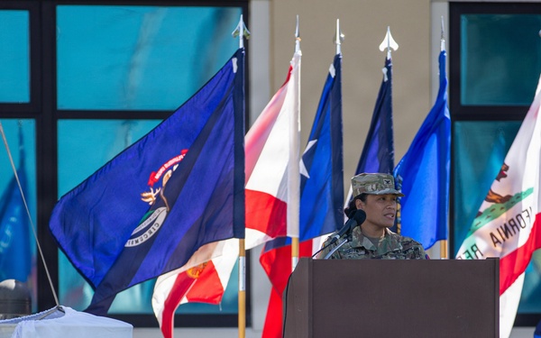 A military service member in camouflage uniform and patrol cap stands at a wooden podium with a microphone, appearing to deliver a speech at an outdoor military ceremony. Multiple U.S. state flags are displayed prominently in the background, including identifiable flags of Louisiana, Texas, and California. In the foreground, an out-of-focus military insignia sign is visible on the grass, consistent with a U.S. Army Medical Command or similar medical unit emblem.