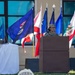 A military service member in camouflage uniform and patrol cap stands at a wooden podium with a microphone, appearing to deliver a speech at an outdoor military ceremony. Multiple U.S. state flags are displayed prominently in the background, including identifiable flags of Louisiana, Texas, and California. In the foreground, an out-of-focus military insignia sign is visible on the grass, consistent with a U.S. Army Medical Command or similar medical unit emblem.