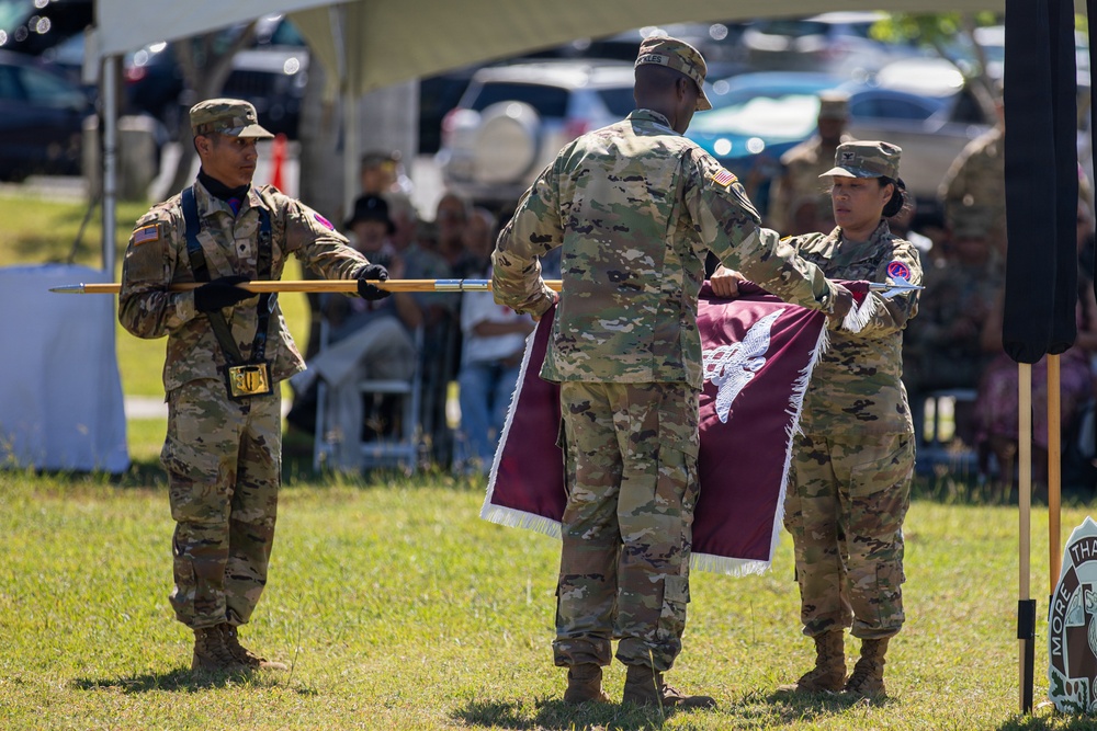 Three U.S. Army soldiers in OCP (Operational Camouflage Pattern) uniforms are performing a military colors ceremony on a grass field. Two soldiers are handling a maroon/burgundy guidon or unit flag with fringe, while a third soldier holds a ceremonial staff or flagpole with black gloves. An audience is seated under a tent canopy in the background, indicating a formal military ceremony such as a change of command or activation/inactivation.