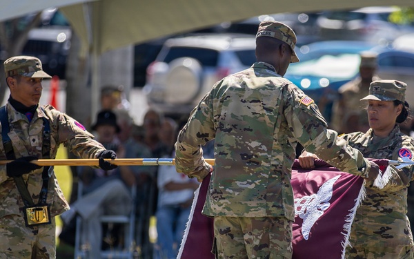 Three U.S. Army soldiers in OCP (Operational Camouflage Pattern) uniforms are performing a military colors ceremony on a grass field. Two soldiers are handling a maroon/burgundy guidon or unit flag with fringe, while a third soldier holds a ceremonial staff or flagpole with black gloves. An audience is seated under a tent canopy in the background, indicating a formal military ceremony such as a change of command or activation/inactivation.