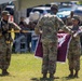 Three U.S. Army soldiers in OCP (Operational Camouflage Pattern) uniforms are performing a military colors ceremony on a grass field. Two soldiers are handling a maroon/burgundy guidon or unit flag with fringe, while a third soldier holds a ceremonial staff or flagpole with black gloves. An audience is seated under a tent canopy in the background, indicating a formal military ceremony such as a change of command or activation/inactivation.