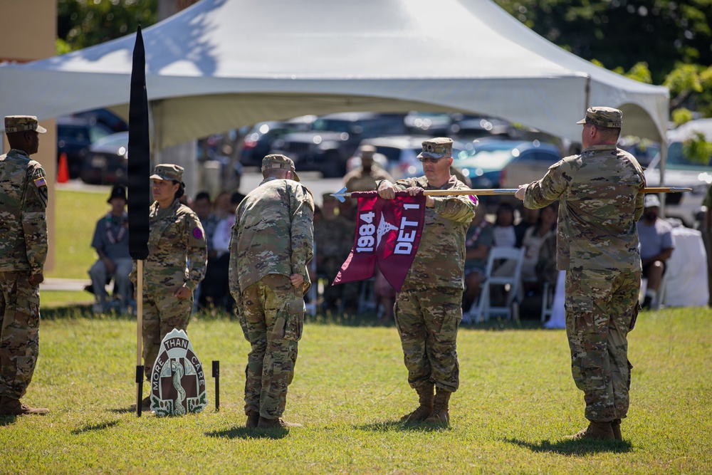 A U.S. Army military ceremony is taking place outdoors on a grass field, likely a unit activation, inactivation, or change of command ceremony. Soldiers in OCP (Operational Camouflage Pattern) uniforms are handling a maroon guidon/flag bearing the text '1984' and 'DET 1', while a unit crest reading 'MORE THAN CARE' with a medical emblem is visible on the ground. An audience of military personnel and civilians is seated under a white tent canopy in the background.