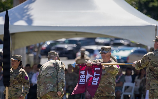 A U.S. Army military ceremony is taking place outdoors on a grass field, likely a unit activation, inactivation, or change of command ceremony. Soldiers in OCP (Operational Camouflage Pattern) uniforms are handling a maroon guidon/flag bearing the text '1984' and 'DET 1', while a unit crest reading 'MORE THAN CARE' with a medical emblem is visible on the ground. An audience of military personnel and civilians is seated under a white tent canopy in the background.