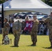 A U.S. Army military ceremony is taking place outdoors on a grass field, likely a unit activation, inactivation, or change of command ceremony. Soldiers in OCP (Operational Camouflage Pattern) uniforms are handling a maroon guidon/flag bearing the text '1984' and 'DET 1', while a unit crest reading 'MORE THAN CARE' with a medical emblem is visible on the ground. An audience of military personnel and civilians is seated under a white tent canopy in the background.
