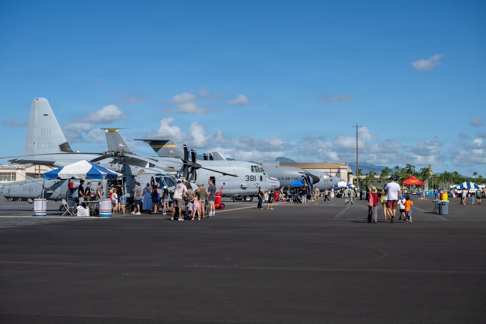 An outdoor military air show or open house event on a tarmac, featuring U.S. Navy and U.S. Air Force aircraft on static display. Civilians including families with children are walking around and inspecting the aircraft up close. The setting appears to be a tropical military airbase, likely in Hawaii based on the mountainous backdrop and palm trees.