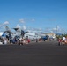 An outdoor military air show or open house event on a tarmac, featuring U.S. Navy and U.S. Air Force aircraft on static display. Civilians including families with children are walking around and inspecting the aircraft up close. The setting appears to be a tropical military airbase, likely in Hawaii based on the mountainous backdrop and palm trees.