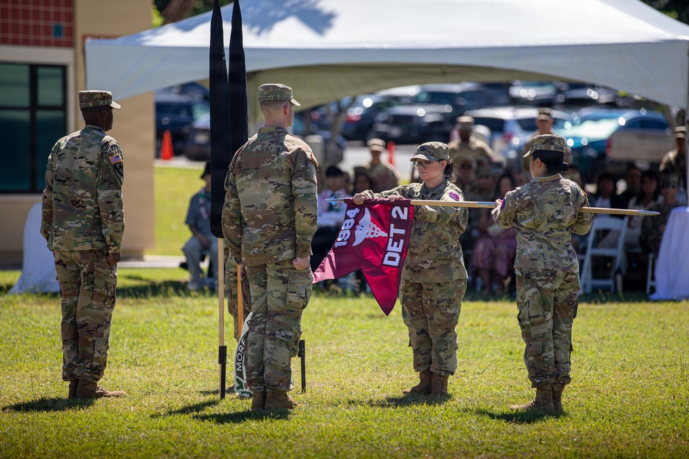 A military ceremony is taking place outdoors on a grass field, with soldiers in U.S. Army OCP (Operational Camouflage Pattern) uniforms participating in what appears to be a unit activation, inactivation, or change of command ceremony. Two soldiers are unfurling or casing a maroon guidon flag marked 'DET 2' and '1984', while other soldiers stand at attention nearby. A seated audience is visible in the background under a white tent canopy.
