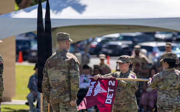 A military ceremony is taking place outdoors on a grass field, with soldiers in U.S. Army OCP (Operational Camouflage Pattern) uniforms participating in what appears to be a unit activation, inactivation, or change of command ceremony. Two soldiers are unfurling or casing a maroon guidon flag marked 'DET 2' and '1984', while other soldiers stand at attention nearby. A seated audience is visible in the background under a white tent canopy.