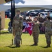 A military ceremony is taking place outdoors on a grass field, with soldiers in U.S. Army OCP (Operational Camouflage Pattern) uniforms participating in what appears to be a unit activation, inactivation, or change of command ceremony. Two soldiers are unfurling or casing a maroon guidon flag marked 'DET 2' and '1984', while other soldiers stand at attention nearby. A seated audience is visible in the background under a white tent canopy.