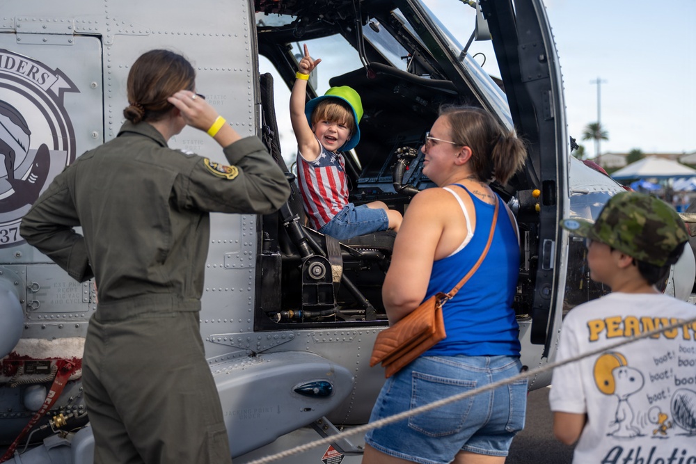 A young child wearing an American flag shirt and green hat sits excitedly in the cockpit of a military helicopter, raising their hand in a peace/victory sign. A woman in a blue tank top stands nearby watching, while a military personnel in an olive flight suit photographs the moment. A second child wearing a Snoopy/Peanuts shirt is partially visible in the foreground.