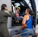 A young child wearing an American flag shirt and green hat sits excitedly in the cockpit of a military helicopter, raising their hand in a peace/victory sign. A woman in a blue tank top stands nearby watching, while a military personnel in an olive flight suit photographs the moment. A second child wearing a Snoopy/Peanuts shirt is partially visible in the foreground.