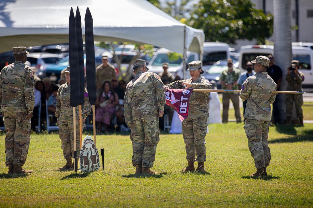 A U.S. Army military ceremony is taking place outdoors on a grass field, likely a casing or uncasing of colors ceremony. Soldiers in Army Combat Uniforms (ACUs) are handling a maroon guidon flag marked 'DET 3' while other personnel stand in formation. A unit insignia shield reading 'MORE THAN CARE' is visible on the ground, suggesting a medical unit affiliation.