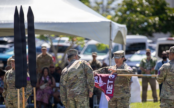 A U.S. Army military ceremony is taking place outdoors on a grass field, likely a casing or uncasing of colors ceremony. Soldiers in Army Combat Uniforms (ACUs) are handling a maroon guidon flag marked 'DET 3' while other personnel stand in formation. A unit insignia shield reading 'MORE THAN CARE' is visible on the ground, suggesting a medical unit affiliation.
