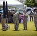 A U.S. Army military ceremony is taking place outdoors on a grass field, likely a casing or uncasing of colors ceremony. Soldiers in Army Combat Uniforms (ACUs) are handling a maroon guidon flag marked 'DET 3' while other personnel stand in formation. A unit insignia shield reading 'MORE THAN CARE' is visible on the ground, suggesting a medical unit affiliation.