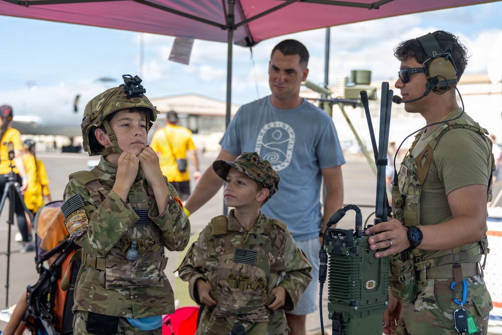 Two children dressed in military camouflage uniforms interact with an active-duty service member at what appears to be a military air show or open house event. The older child is adjusting a tactical helmet with a night-vision mount, while a younger child in a patrol cap looks on. An adult civilian, likely a parent, stands in the background under a red canopy tent.