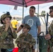Two children dressed in military camouflage uniforms interact with an active-duty service member at what appears to be a military air show or open house event. The older child is adjusting a tactical helmet with a night-vision mount, while a younger child in a patrol cap looks on. An adult civilian, likely a parent, stands in the background under a red canopy tent.
