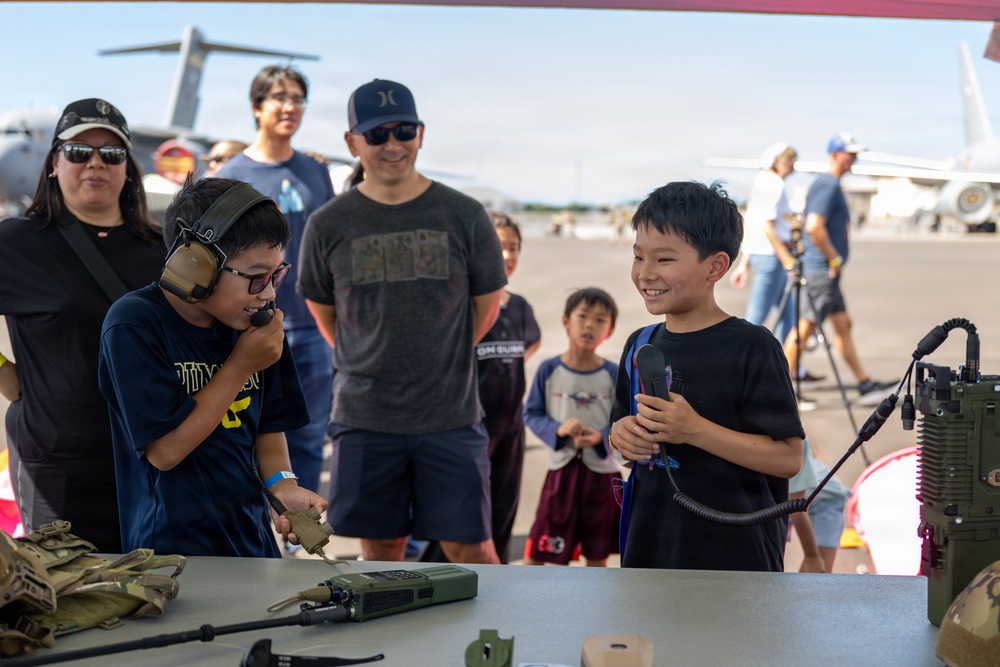 Two young boys interact with military communication equipment at what appears to be an air show or military open house event. One boy is wearing tactical headphones and speaking into a radio handset, while the other holds a microphone and smiles broadly. A group of adults and children observe in the background near military aircraft on a tarmac.