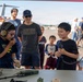 Two young boys interact with military communication equipment at what appears to be an air show or military open house event. One boy is wearing tactical headphones and speaking into a radio handset, while the other holds a microphone and smiles broadly. A group of adults and children observe in the background near military aircraft on a tarmac.
