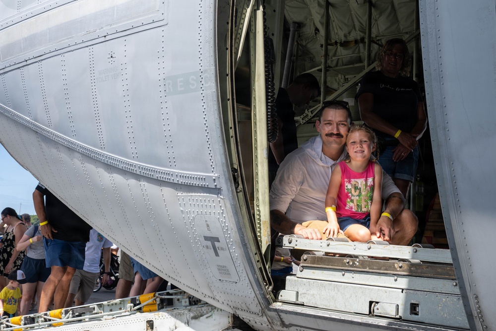 A man with a mustache and a young girl are seated together in the open cargo door or ramp of a large military aircraft, smiling at the camera. The setting appears to be a public airshow or open-house event, with crowds of visitors visible outside the aircraft. Multiple people wearing yellow wristbands are present, suggesting a ticketed or organized public event.