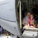 A man with a mustache and a young girl are seated together in the open cargo door or ramp of a large military aircraft, smiling at the camera. The setting appears to be a public airshow or open-house event, with crowds of visitors visible outside the aircraft. Multiple people wearing yellow wristbands are present, suggesting a ticketed or organized public event.