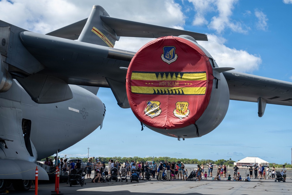 An air show or open house event at Hickam Air Force Base (Joint Base Pearl Harbor-Hickam, Hawaii), featuring a large military transport aircraft with a decorative red engine inlet cover displaying the 'HICKAM' name and military unit insignia. A crowd of civilians including families with strollers are gathered on the tarmac viewing the aircraft. The scene takes place under partly cloudy skies in what appears to be a tropical setting.