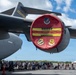 An air show or open house event at Hickam Air Force Base (Joint Base Pearl Harbor-Hickam, Hawaii), featuring a large military transport aircraft with a decorative red engine inlet cover displaying the 'HICKAM' name and military unit insignia. A crowd of civilians including families with strollers are gathered on the tarmac viewing the aircraft. The scene takes place under partly cloudy skies in what appears to be a tropical setting.