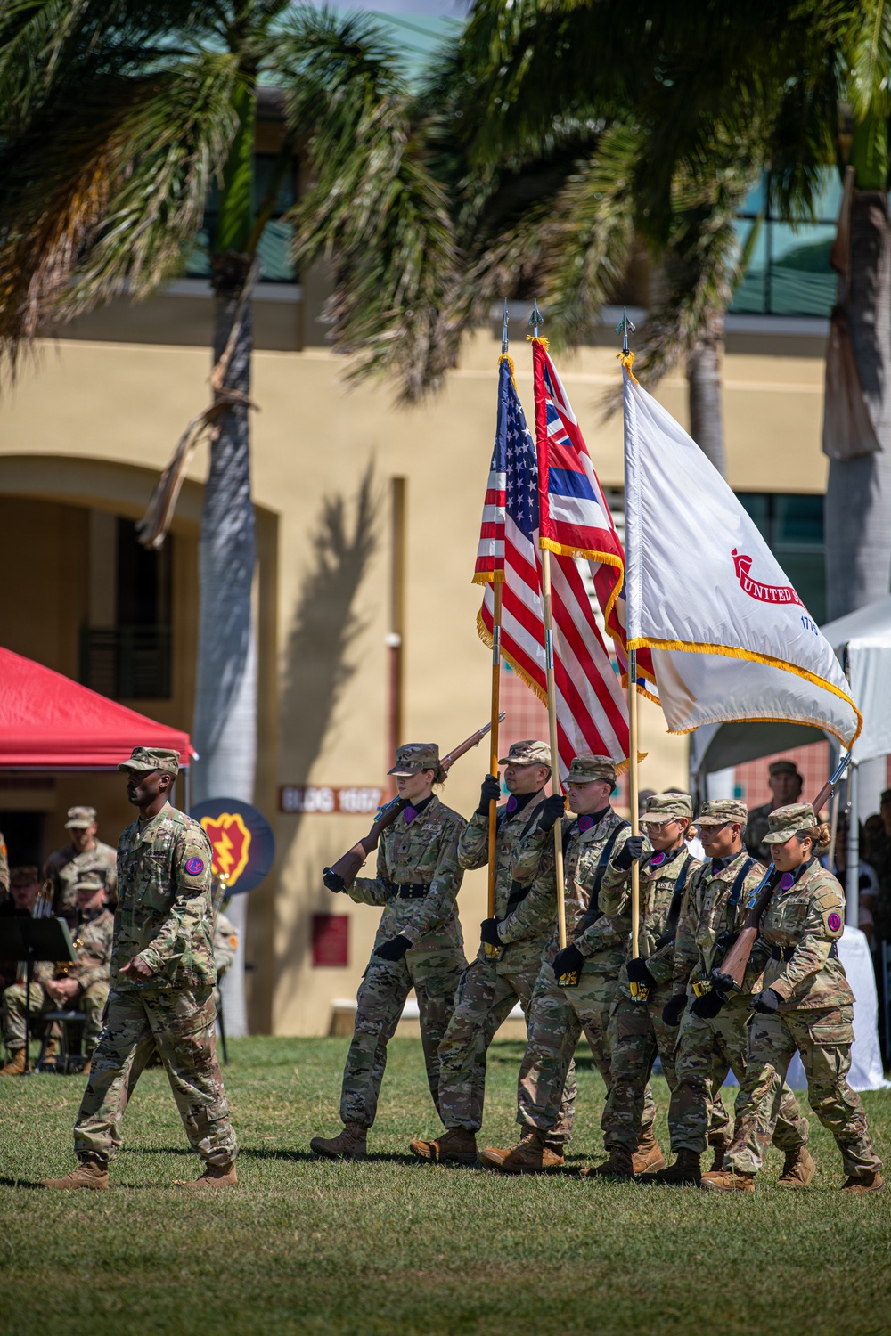 A military color guard ceremony is taking place outdoors on a grassy parade field, likely at a U.S. Army installation in Hawaii. Personnel in Army Combat Uniforms (ACU/OCP pattern) are carrying multiple flags including the U.S. national flag, the Hawaii state flag, and a white unit guidon. The setting includes palm trees, a tan building with 'BLDG 1002' visible, and a unit insignia shield in the background consistent with the 25th Infantry Division.