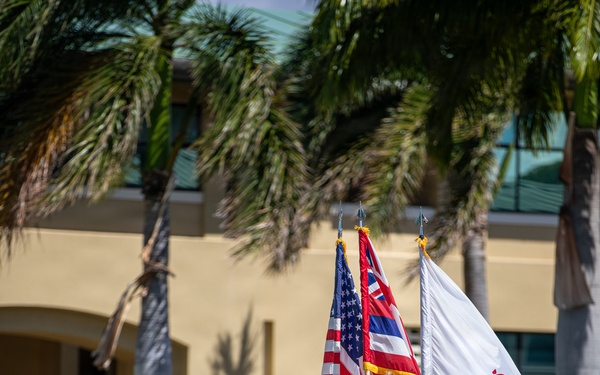 A military color guard ceremony is taking place outdoors on a grassy parade field, likely at a U.S. Army installation in Hawaii. Personnel in Army Combat Uniforms (ACU/OCP pattern) are carrying multiple flags including the U.S. national flag, the Hawaii state flag, and a white unit guidon. The setting includes palm trees, a tan building with 'BLDG 1002' visible, and a unit insignia shield in the background consistent with the 25th Infantry Division.