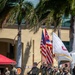 A military color guard ceremony is taking place outdoors on a grassy parade field, likely at a U.S. Army installation in Hawaii. Personnel in Army Combat Uniforms (ACU/OCP pattern) are carrying multiple flags including the U.S. national flag, the Hawaii state flag, and a white unit guidon. The setting includes palm trees, a tan building with 'BLDG 1002' visible, and a unit insignia shield in the background consistent with the 25th Infantry Division.