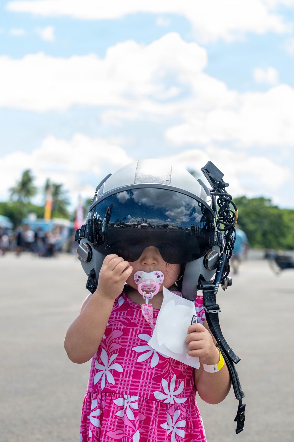 A toddler wearing an oversized military/aviation flight helmet with a dark visor stands outdoors at what appears to be an airshow or military open day event. The child is dressed in a pink floral dress, has a pink pacifier in her mouth, and is holding a white paper item. The helmet is far too large for the child, creating a humorous and endearing visual contrast.